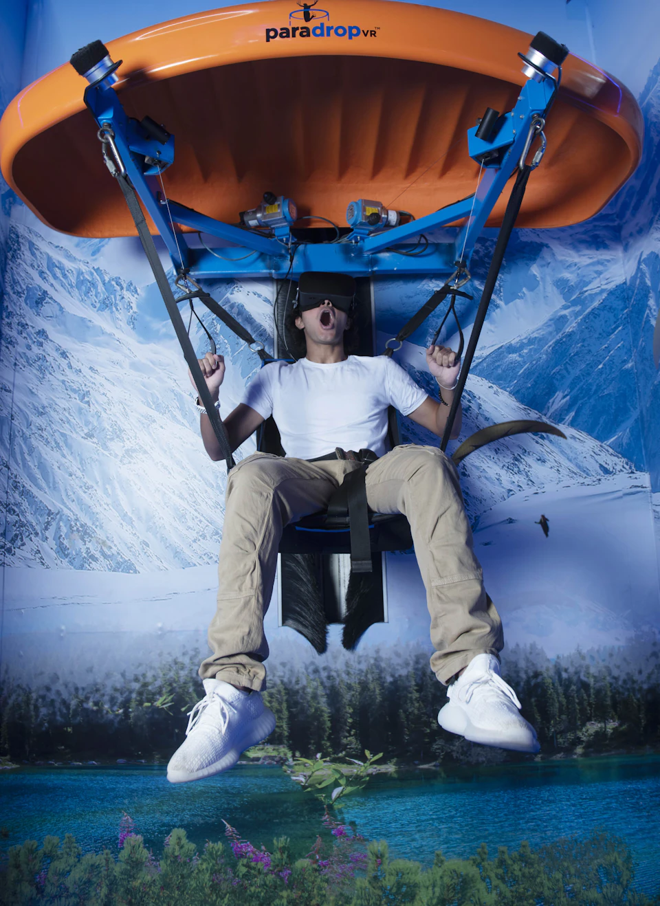 A man wearing a VR headset is seated on a suspended ride against a backdrop of snowy mountains, appearing excited.
