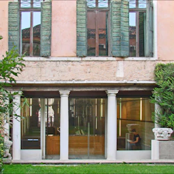 En yttre vy av en byggnad med stora fönster och gröna fönsterluckor, med pelare och frodig grönska framför.