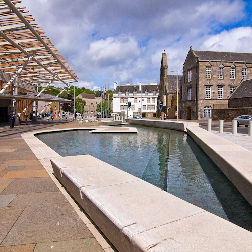 Utomhusscen med en modern byggnad, stenbeläggning och rektangulär vattenfunktion. Historiska byggnader och lyktstolpar i bakgrunden.