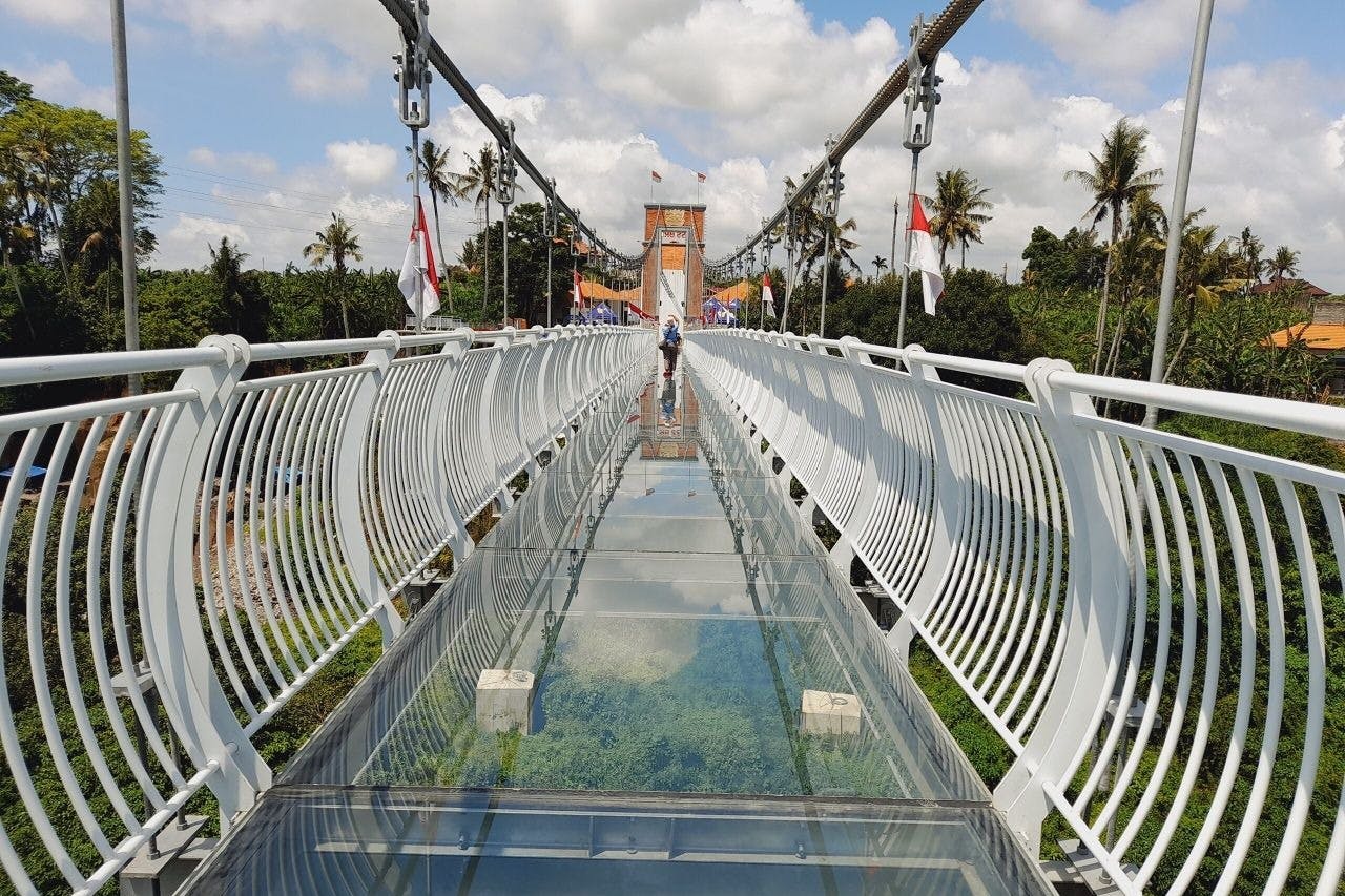 Člověk kráčí po visutém mostě se skleněným dnem a bílým zábradlím, obklopen stromy a vlajkami pod částečně zataženou oblohou.