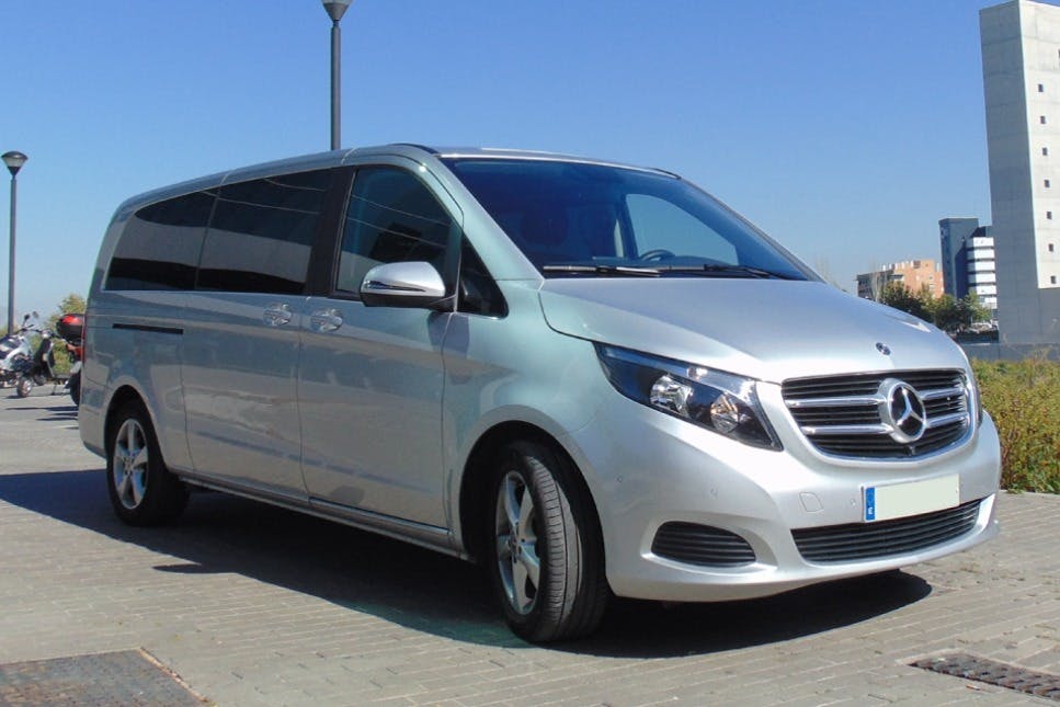 Stříbrná dodávka Mercedes-Benz zaparkovaná na dlážděné ulici s budovami a jasnou oblohou v pozadí.