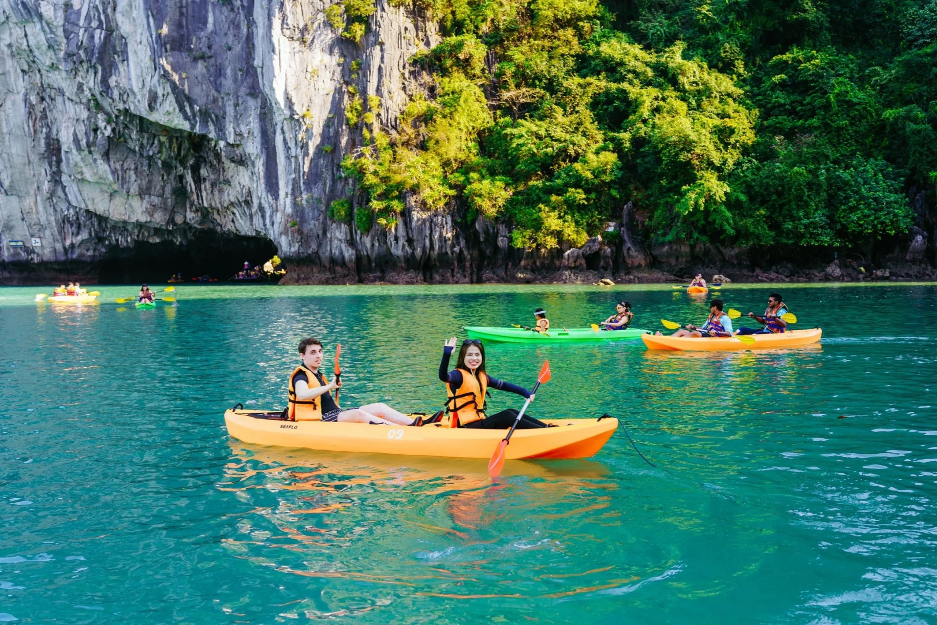 Two people in a yellow kayak paddle on turquoise water near a rocky cliff and greenery, with other kayakers in the background.