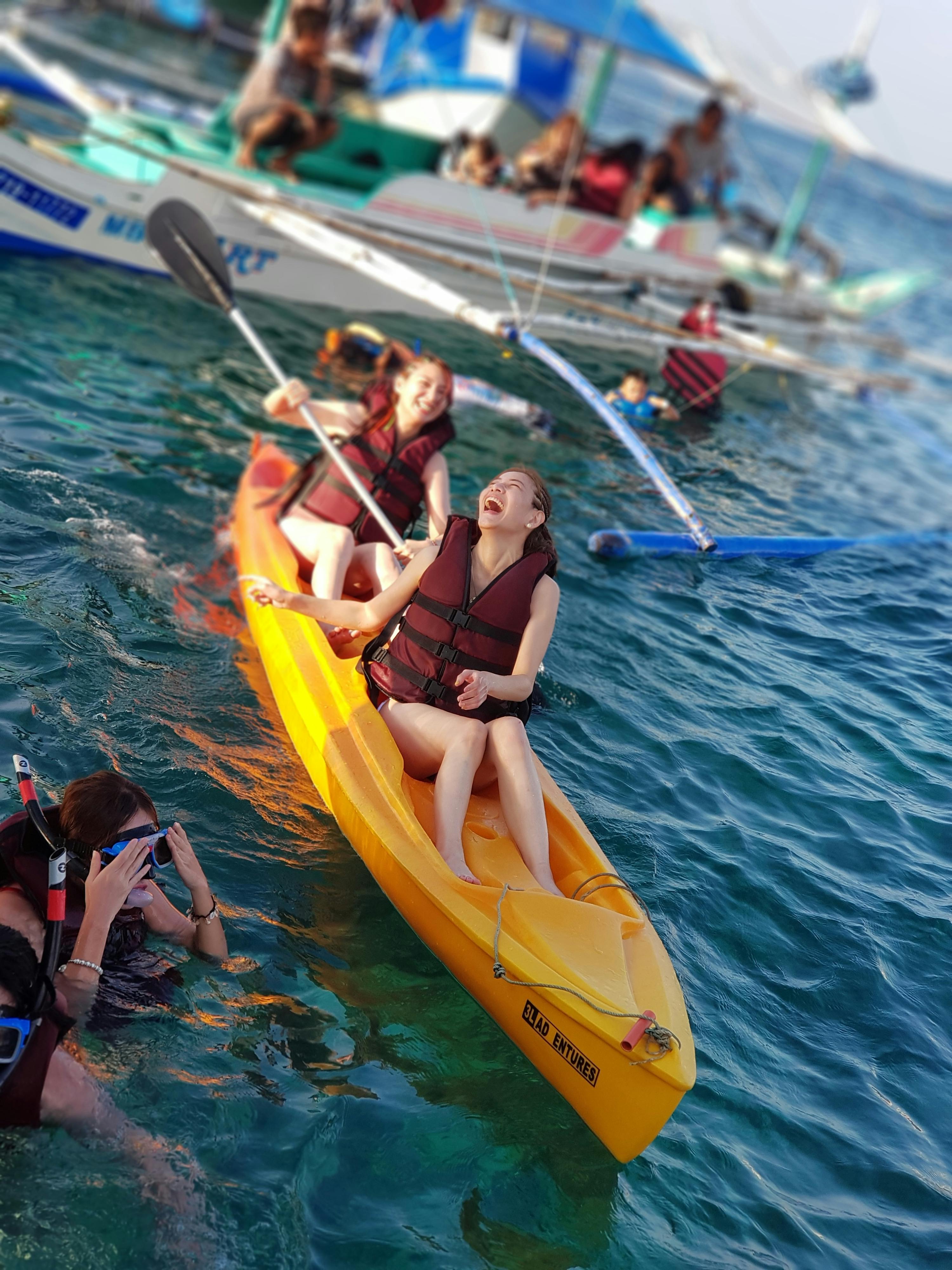 Two people wearing life jackets are enjoying a ride on a yellow kayak in a water body, with boats visible in the background.