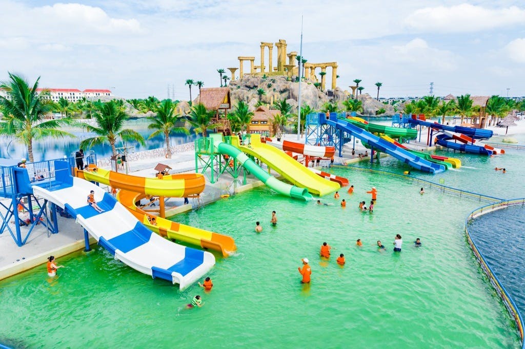 People enjoying colorful water slides in a vibrant water park with a backdrop of palm trees and replica ancient ruins.