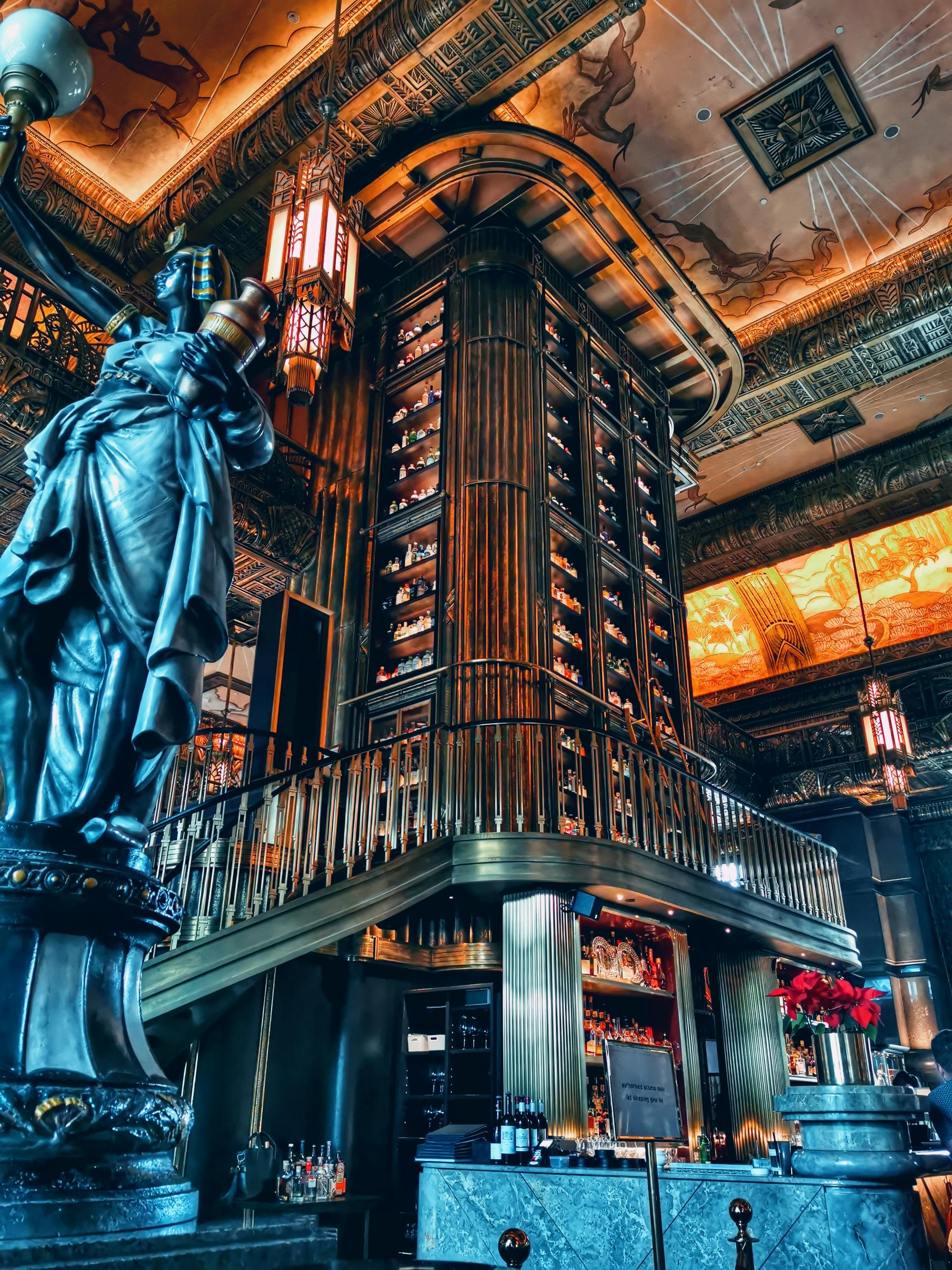 Opulent interior featuring a towering, cylindrical wooden wine rack, intricate ceiling designs, and a statue of a robed figure holding a torch.