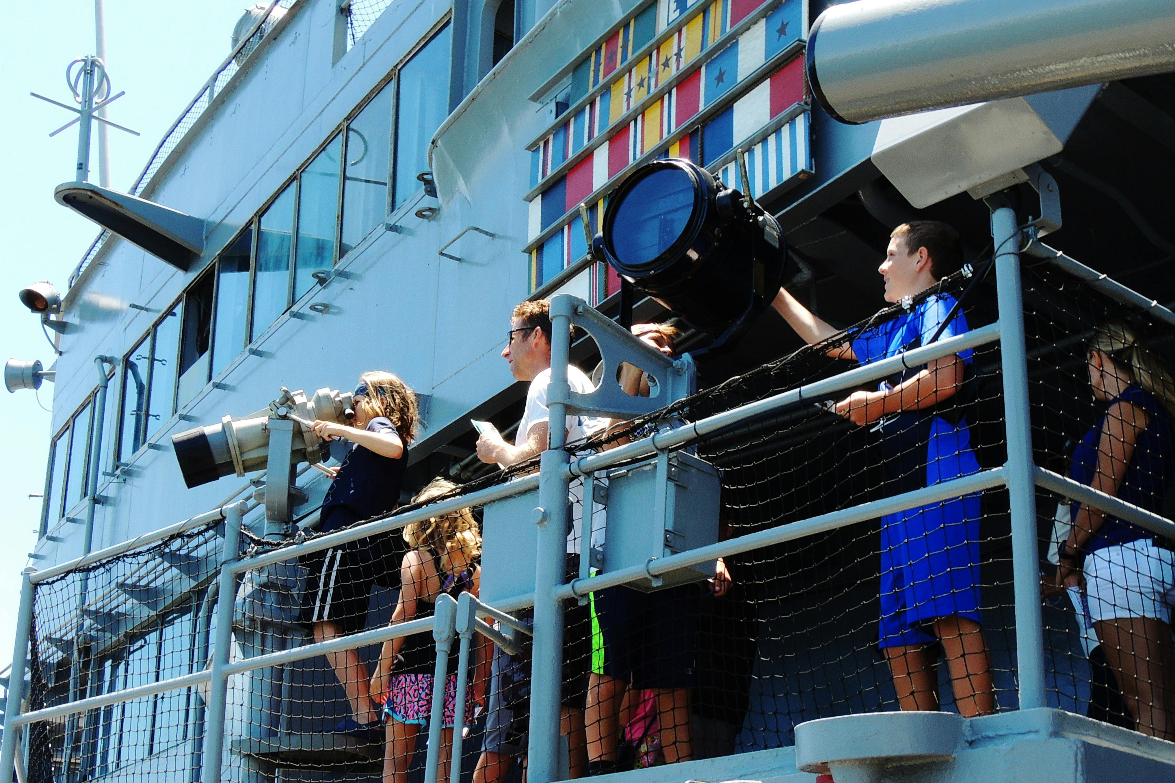 People standing on the deck of a ship near a large light fixture, with colorful flags displayed in the background.