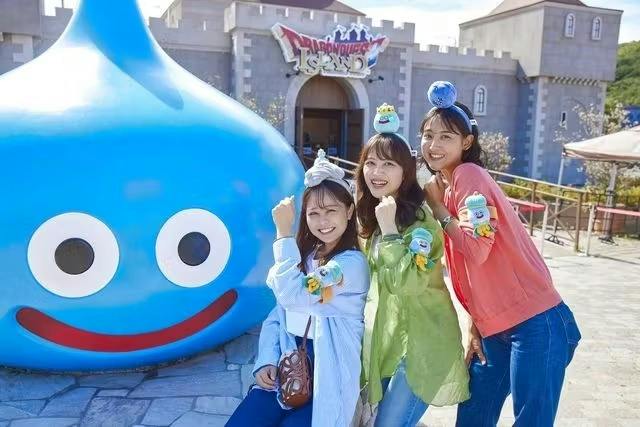 Three smiling women posing in front of a large blue slime statue at a themed attraction, wearing matching headbands and wristbands.