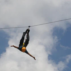 En person med en gul hjälm hänger upp och ner från en zipline mot en bakgrund av blå himmel och moln.