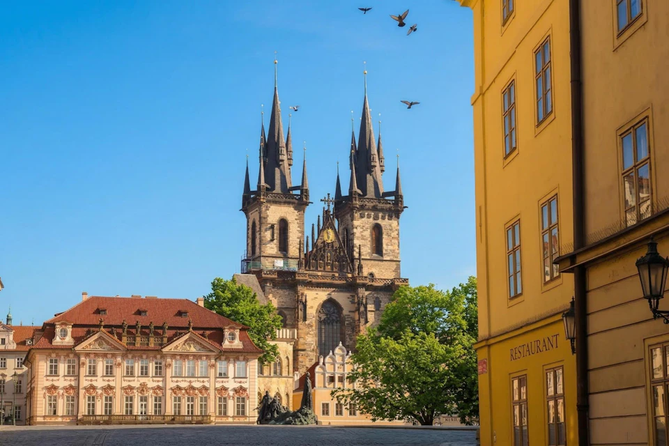 A historic cathedral with spires, surrounded by colorful buildings and trees under a clear blue sky with birds flying.