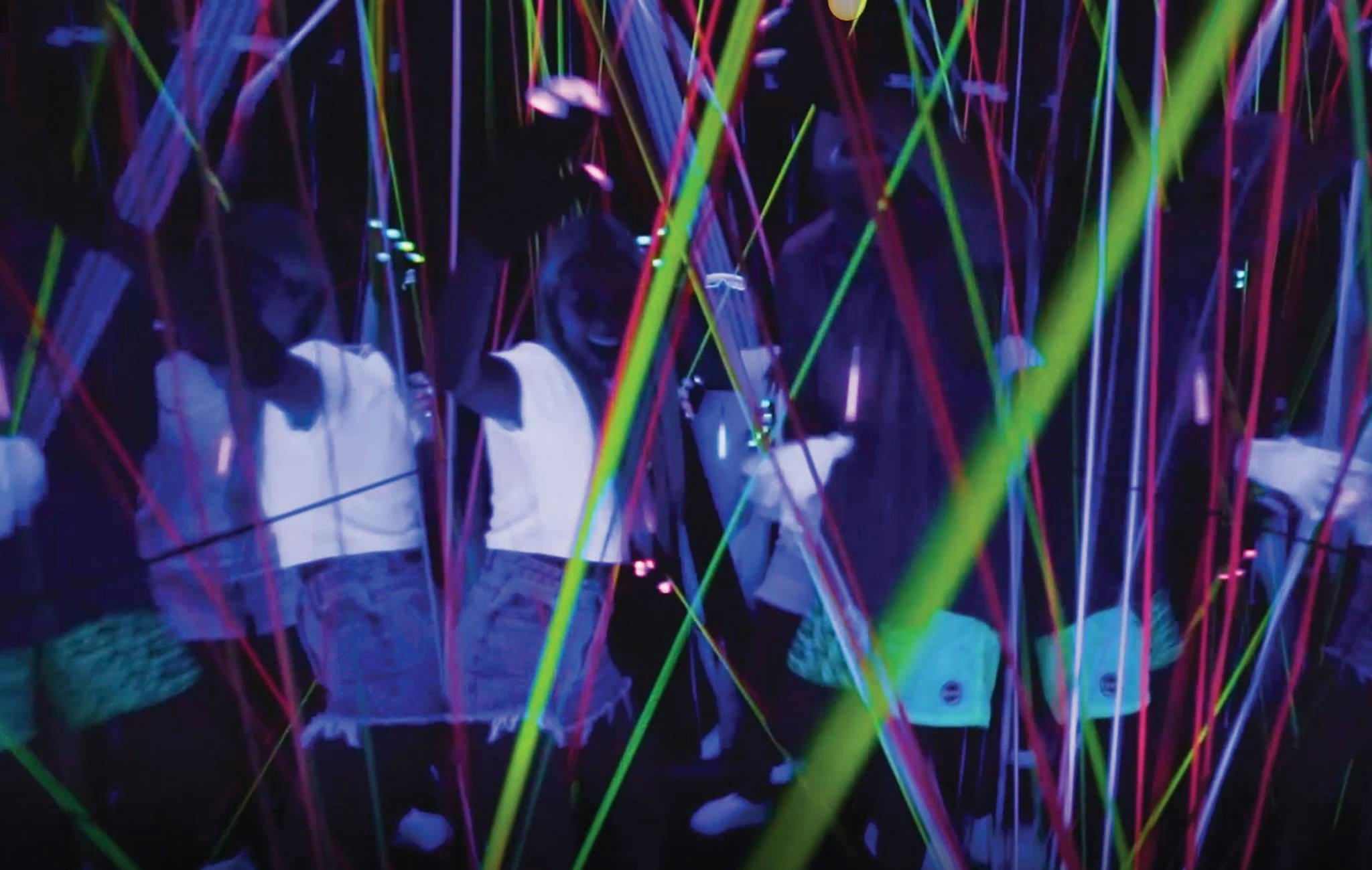 Mensen genieten van een neonverlichte ruimte, met kleurrijke laserstralen en reflecties die een levendige, energieke sfeer creëren.