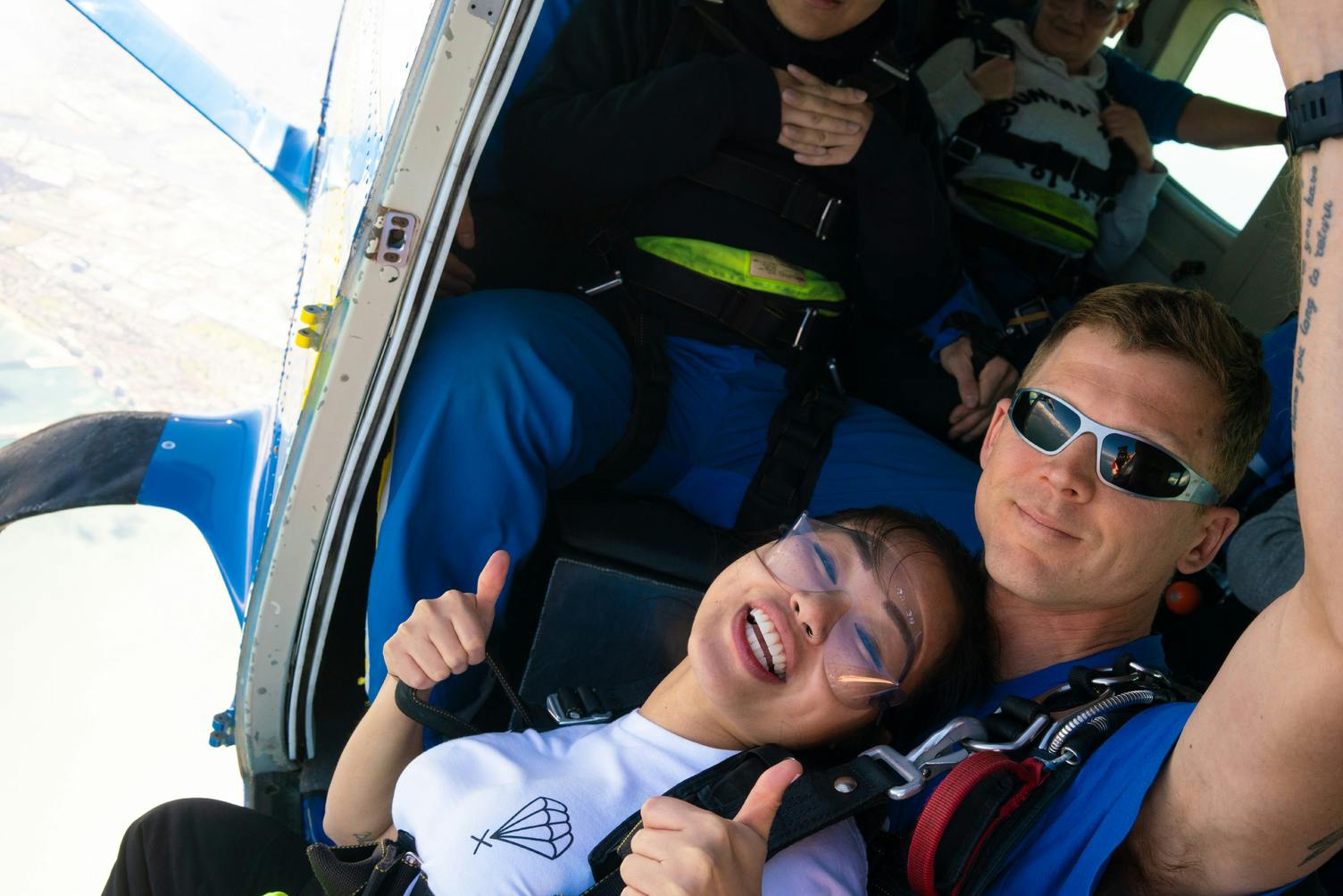 Δύο άνθρωποι με εξοπλισμό ελεύθερης πτώσης δίνουν τον αντίχειρά τους μέσα σε ένα αεροπλάνο, χαμογελώντας. Άλλοι κάθονται στο παρασκήνιο.