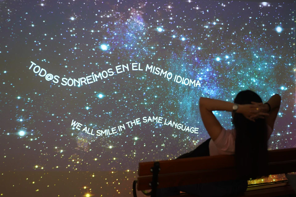 A person sitting on a bench looking at a starry backdrop with text: 'We all smile in the same language.'