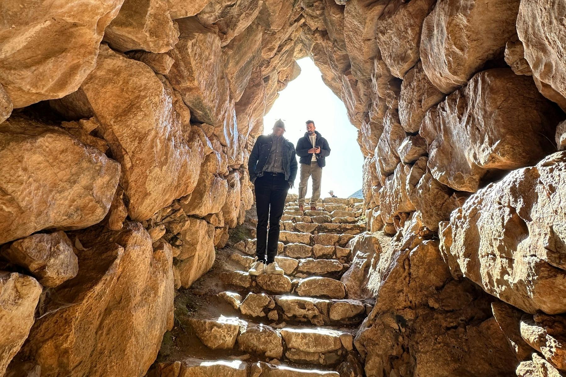 Two visitors walk up stone steps inside a narrow Mycenaean tunnel with large stones on each side.
