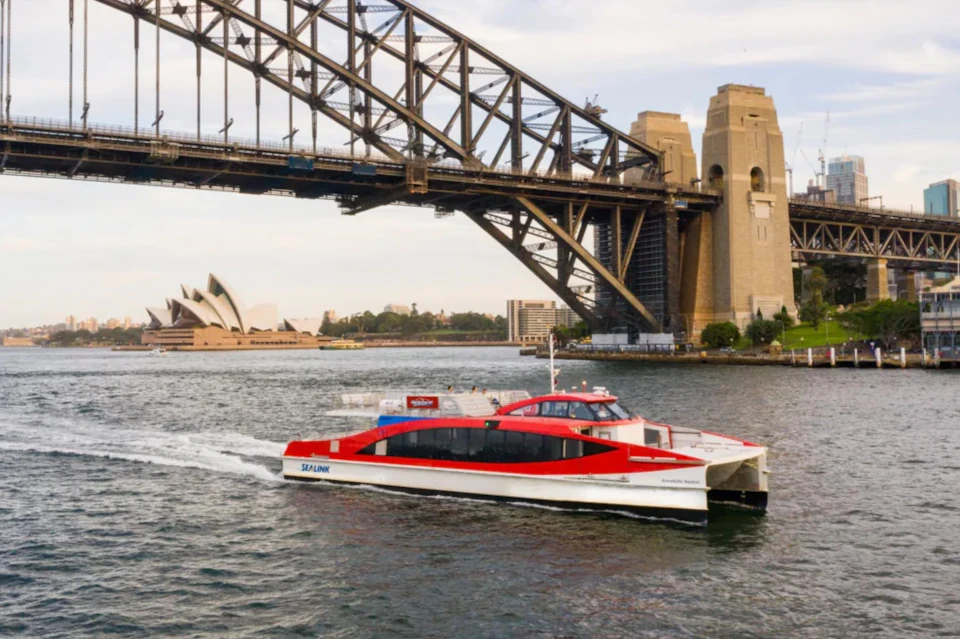 A red and white ferry labeled 'SeaLink' sails under Sydney Harbour Bridge, with the Sydney Opera House visible in the background.