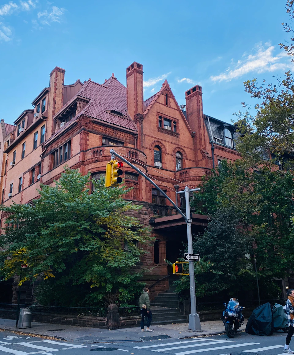 A red brick building with ornate architecture and a steep roof, surrounded by trees, with a streetlight in the foreground.