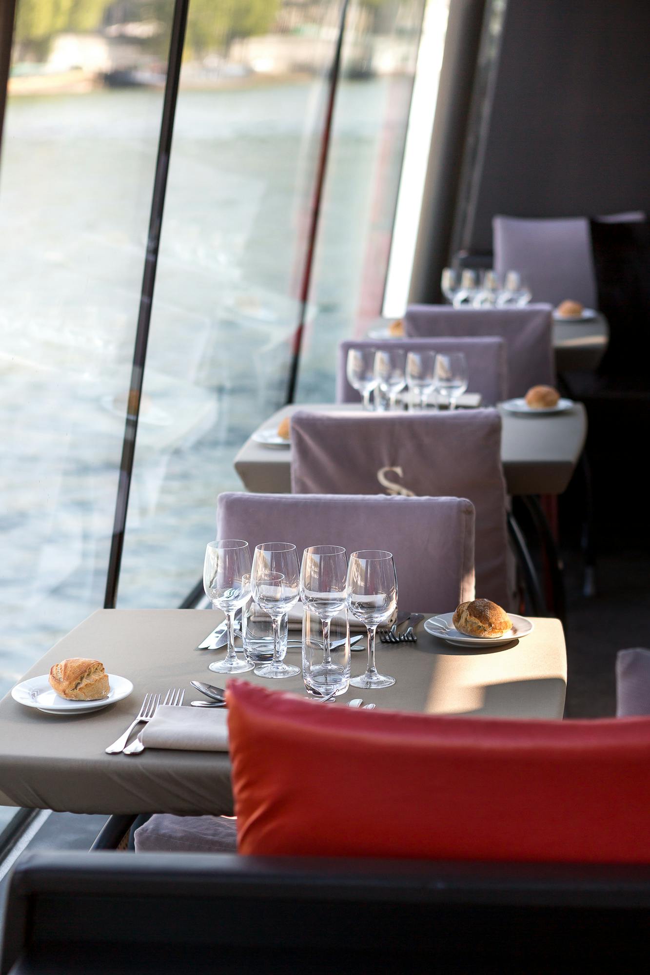 Elegantní restaurační stoly se sklenicemi, příbory a talíři s chlebem, vedle velkých oken s výhledem na vodu.