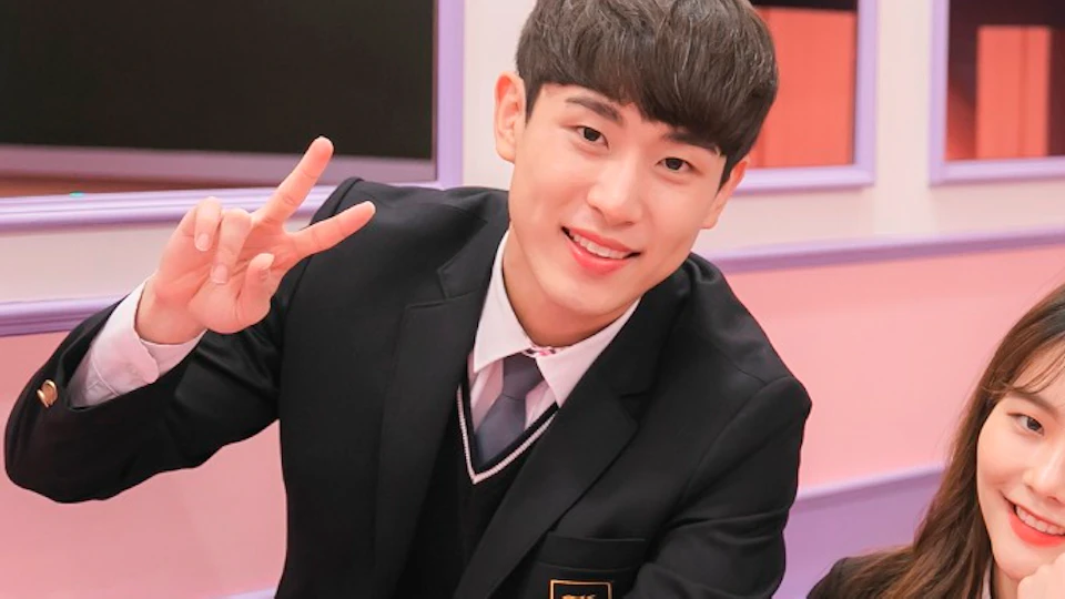Three students in school uniforms sitting and standing in a pastel-colored classroom setting with books and a chalkboard.
