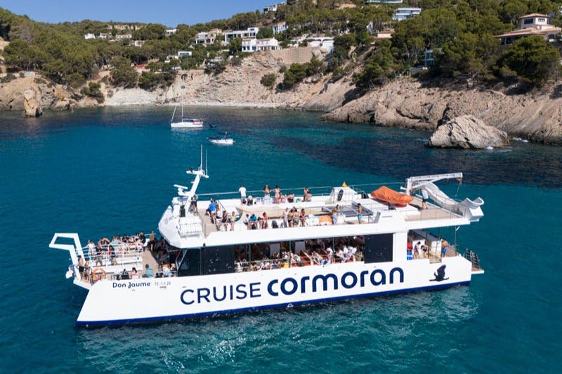 A cruise ship named 'Cormoran' with passengers onboard, sailing in clear blue water near a rocky coast with houses visible.