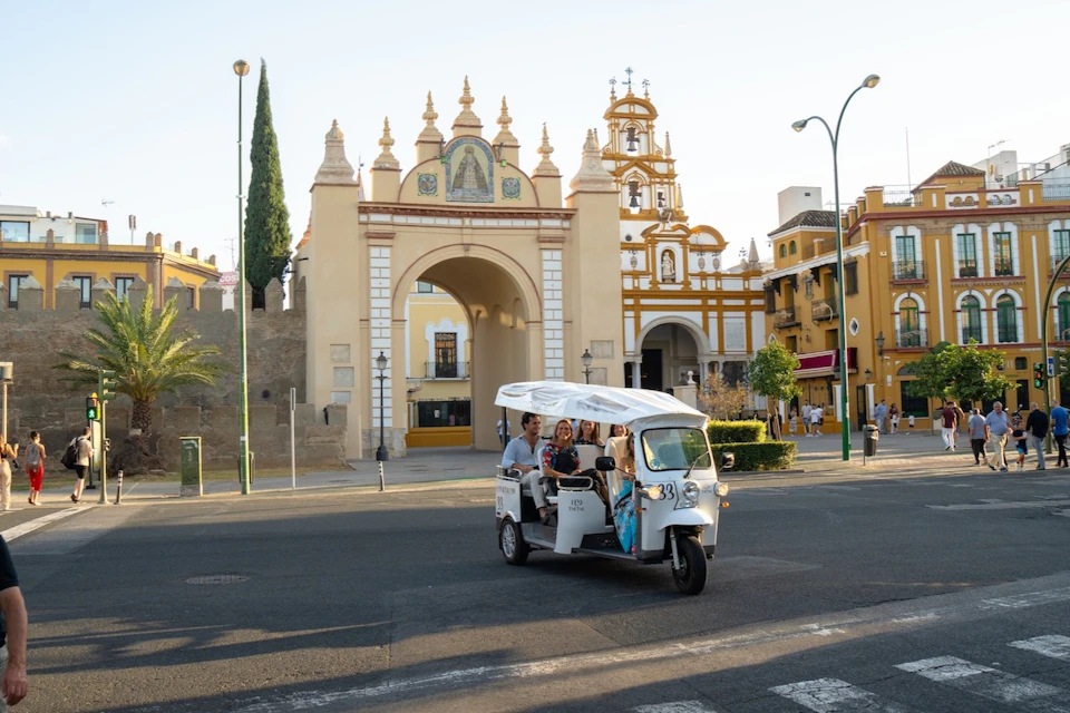 A white tuk-tuk with passengers rides past an ornate archway and church in a sunny plaza.