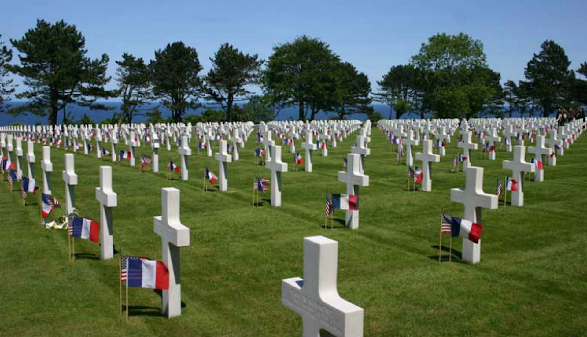 Rows of white crosses in a cemetery with French and American flags, surrounded by trees and under a clear blue sky.