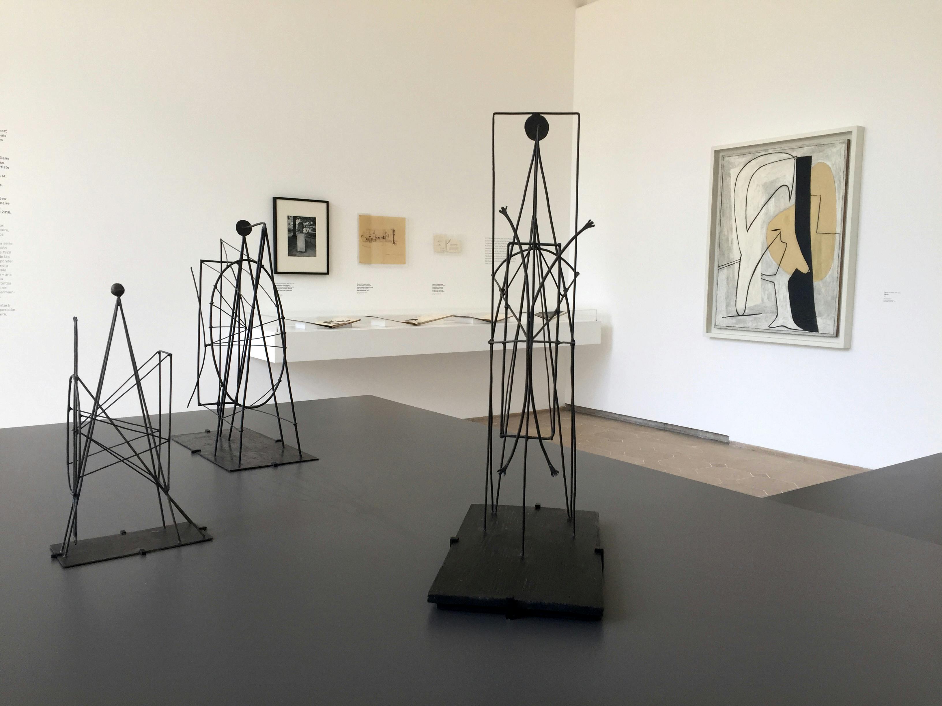 Room with abstract wire sculptures on black bases, drawings on the wall, and a large framed abstract painting.