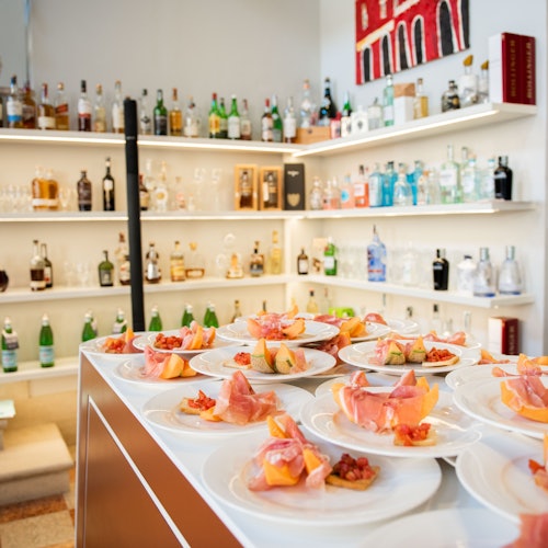 A bar setup with shelves full of various bottles and plates of appetizers with sliced meat and melon on a counter.