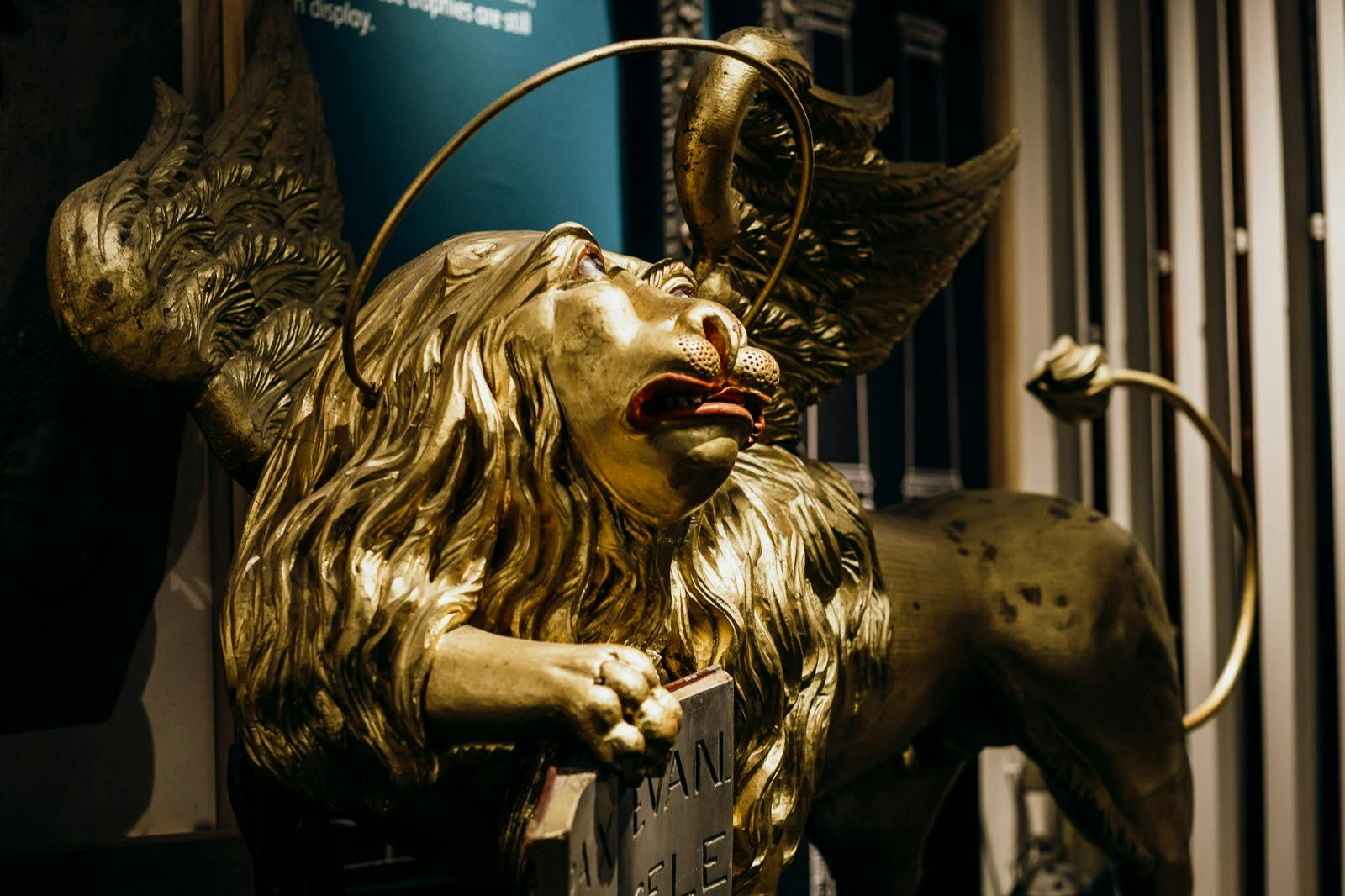 A close-up of a golden lion statue with wings, a halo, and inscriptions on the base, positioned indoors with a dark background.