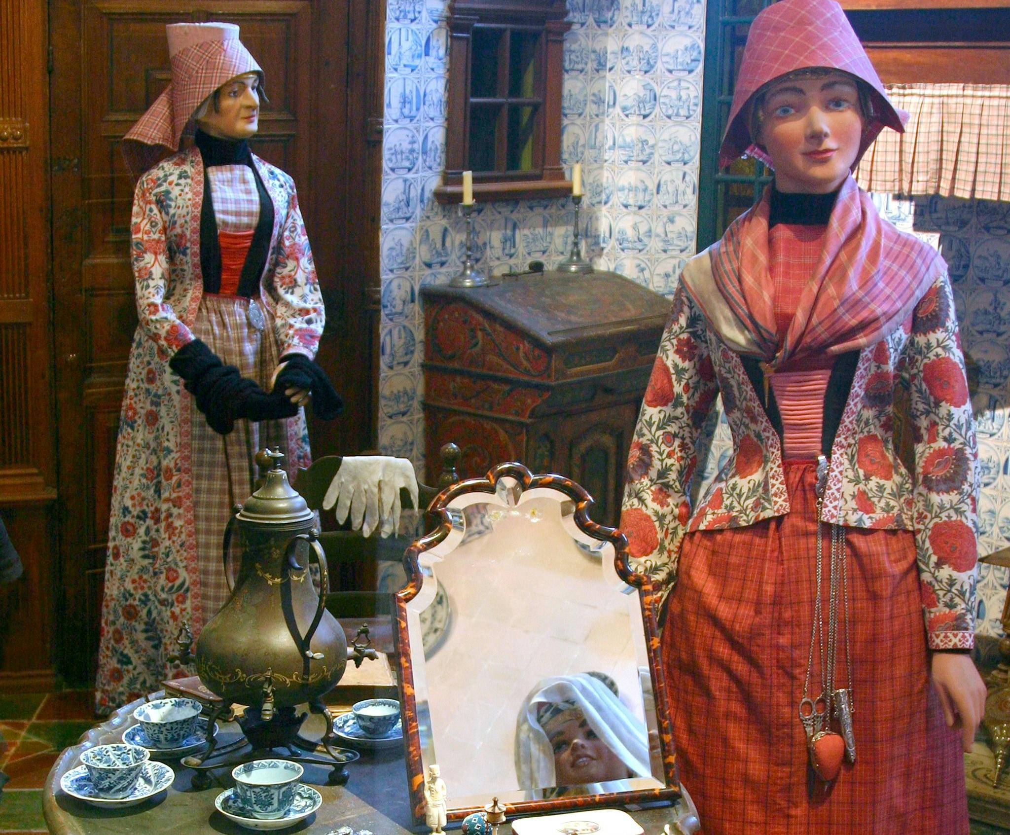 Two mannequins in traditional attire are in a vintage room with patterned walls, a large mirror, and a tea set on a table.