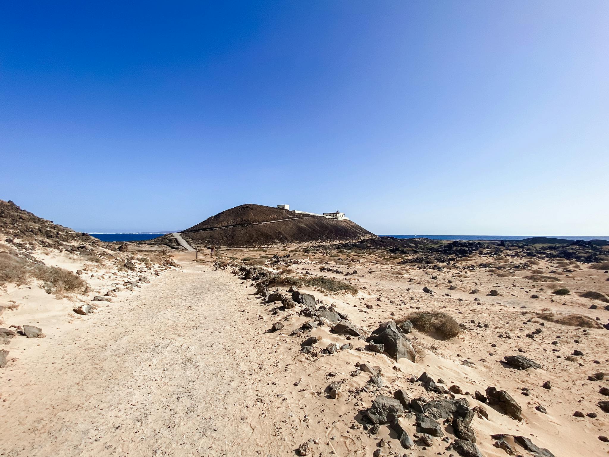 岩だらけの土道は、澄んだ青い空の下にあり、まばらな砂漠の地形に囲まれた、上に建造物がある丘に通じています。