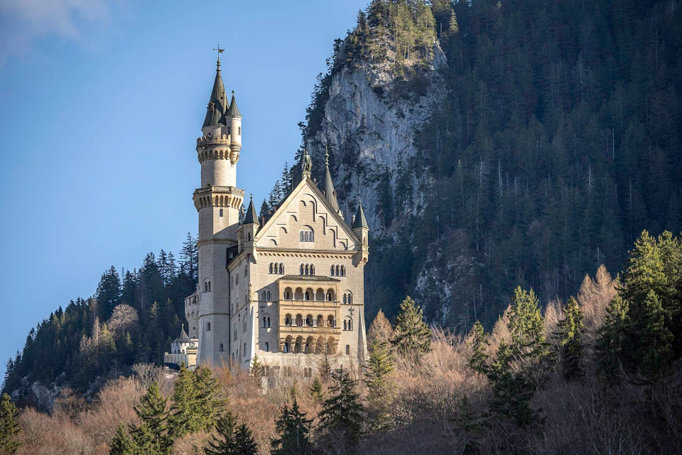 A castle with tall spires set against a forested mountain backdrop under a clear blue sky.