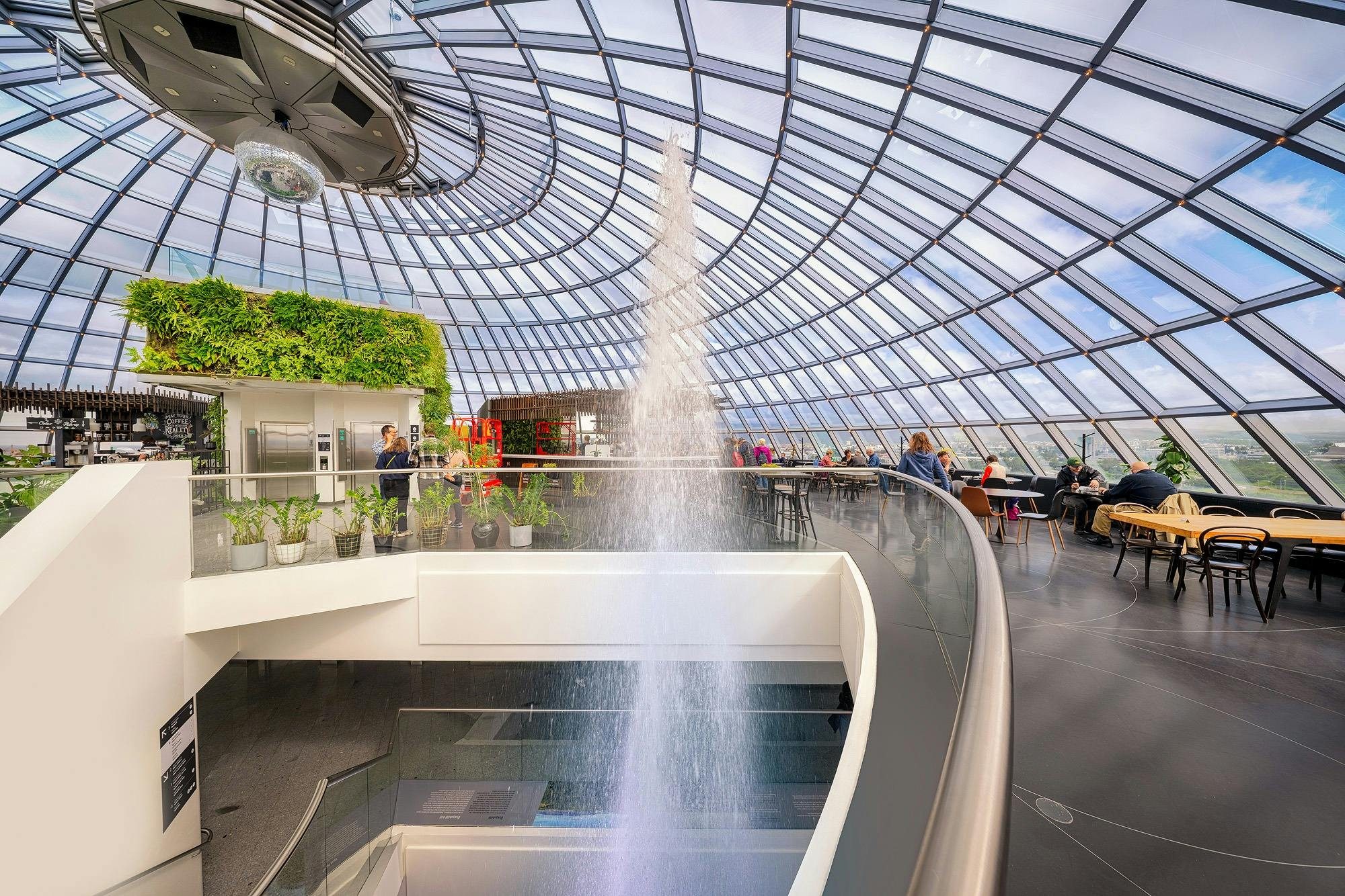 Indendørs atrium med glastag, springvand, planter og folk, der sidder og går rundt.