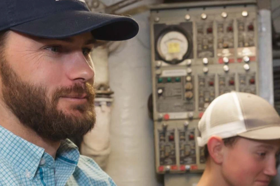 Two men operate machinery in an industrial setting with gauges and wires visible around them.