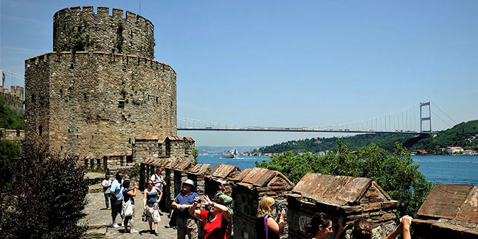 Tourists explore an ancient stone structure with a large tower. In the background, there's a suspension bridge over a body of water.