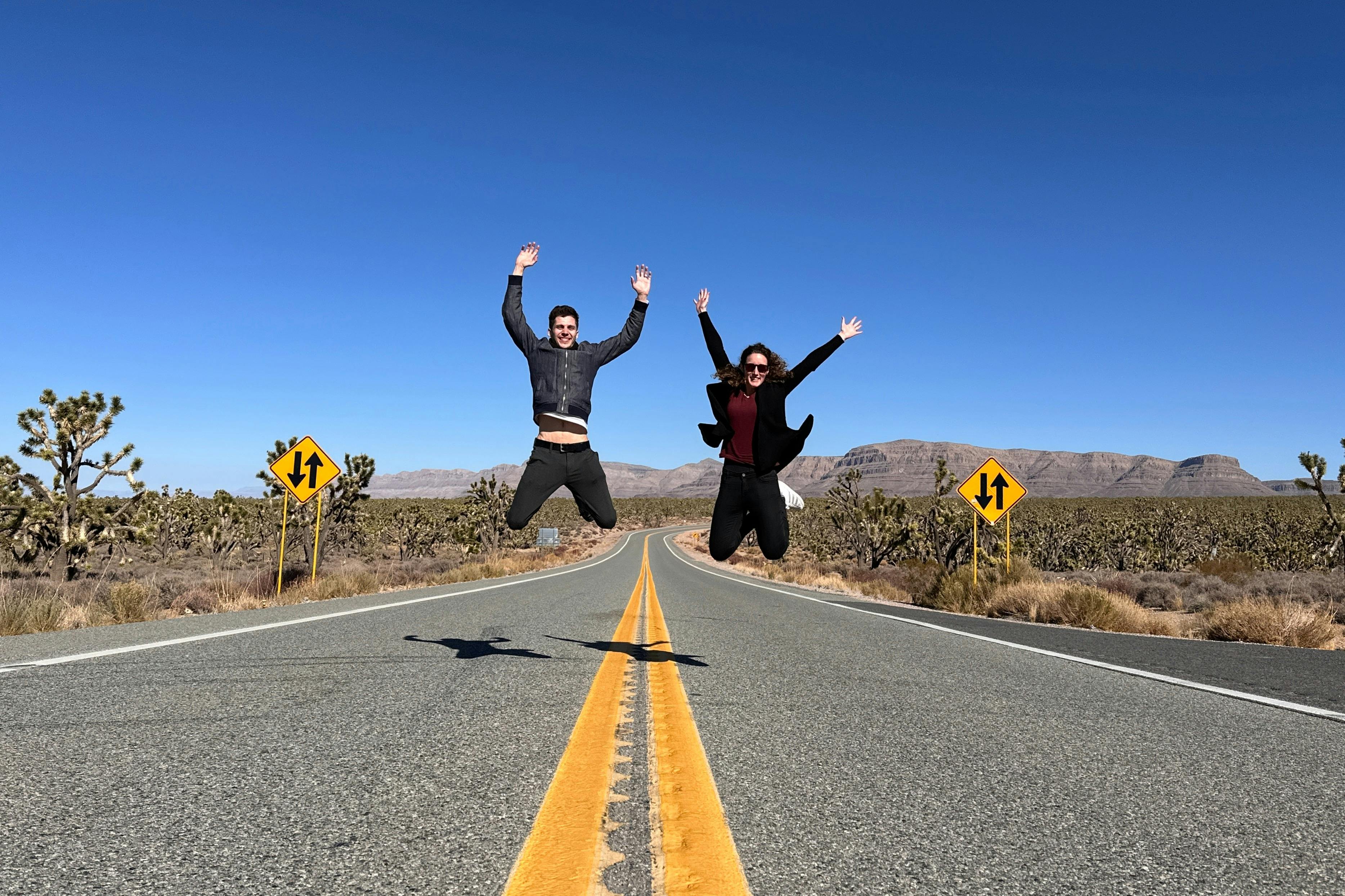 Two people jump with raised arms on an empty road surrounded by desert terrain and traffic signs, under a clear blue sky.
