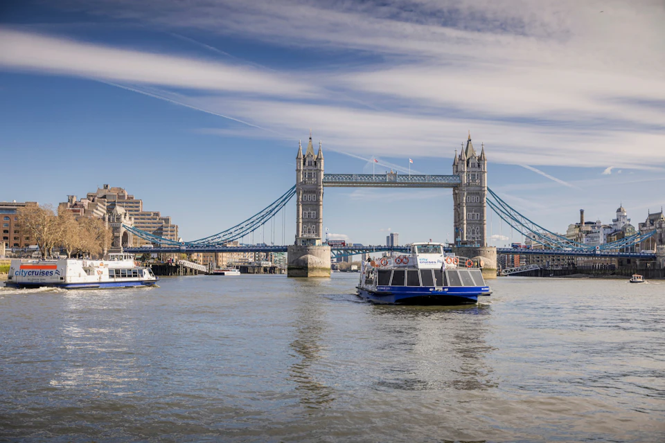 Tower Bridge over the Thames River with boats on the water and a clear sky in the background.