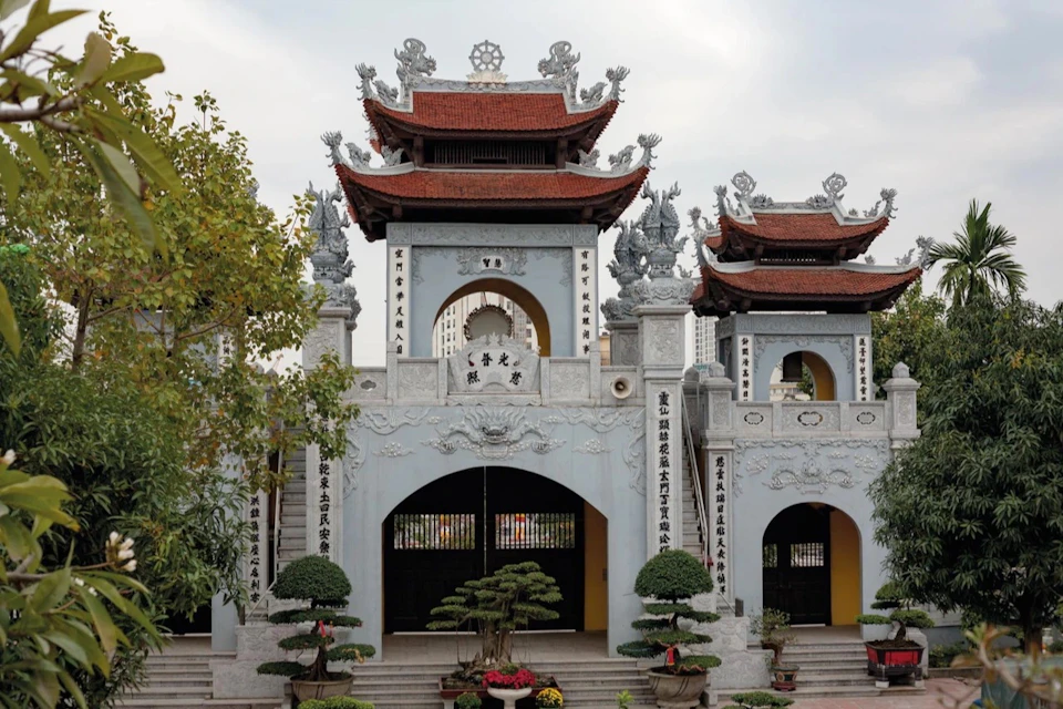A traditional Asian temple entrance with ornate stonework, red-tiled roofs, and bonsai trees in the foreground.