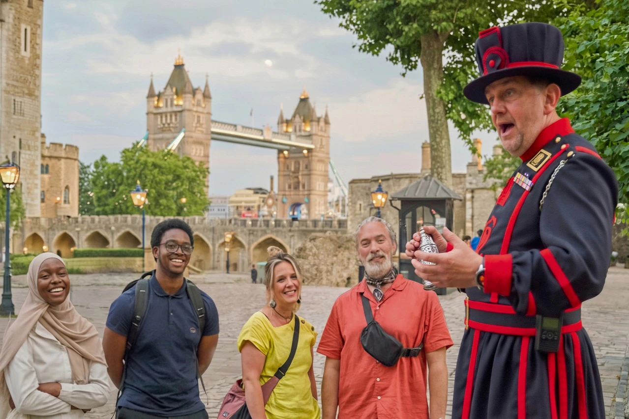 A group of people smiling while a Yeoman Warder speaks to them, with the Tower Bridge visible in the background.