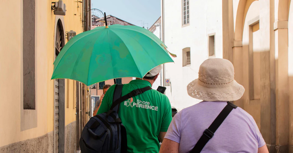 Two people walking down a sunny street, one holding a large green umbrella and wearing a green shirt that says "Shore Experience."