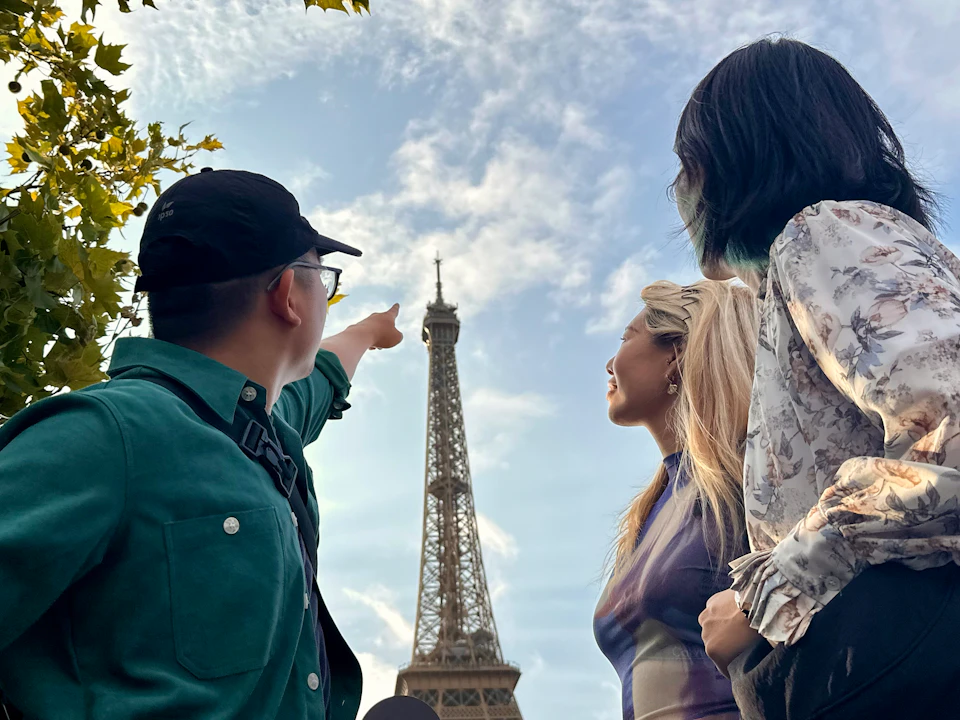 Three people stand near the Eiffel Tower. One points upwards, creating a sense of awe. The sky is partly cloudy.