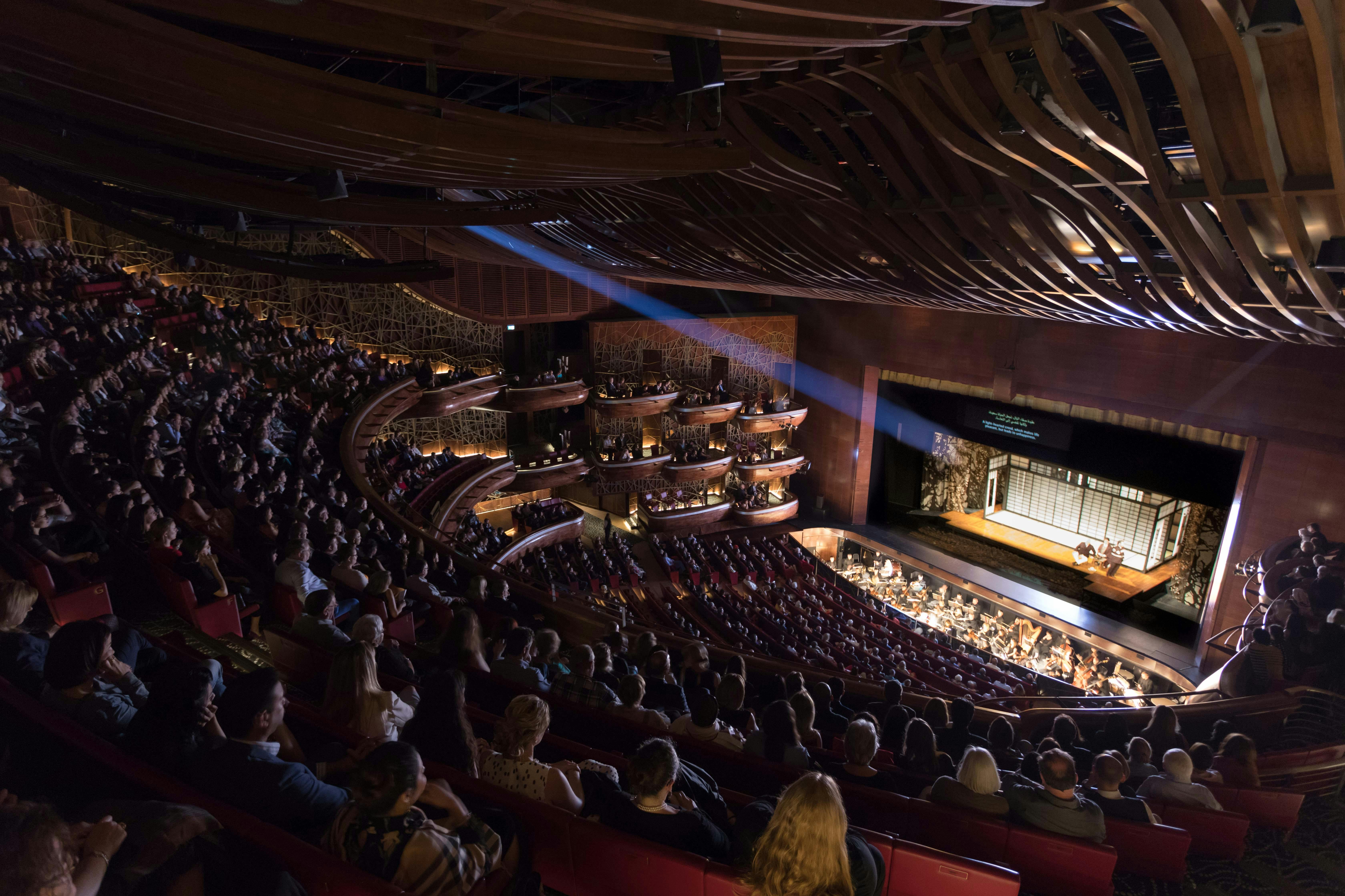 A theater filled with an audience, showing multiple balconies, a stage with performers, and intricate ceiling design.