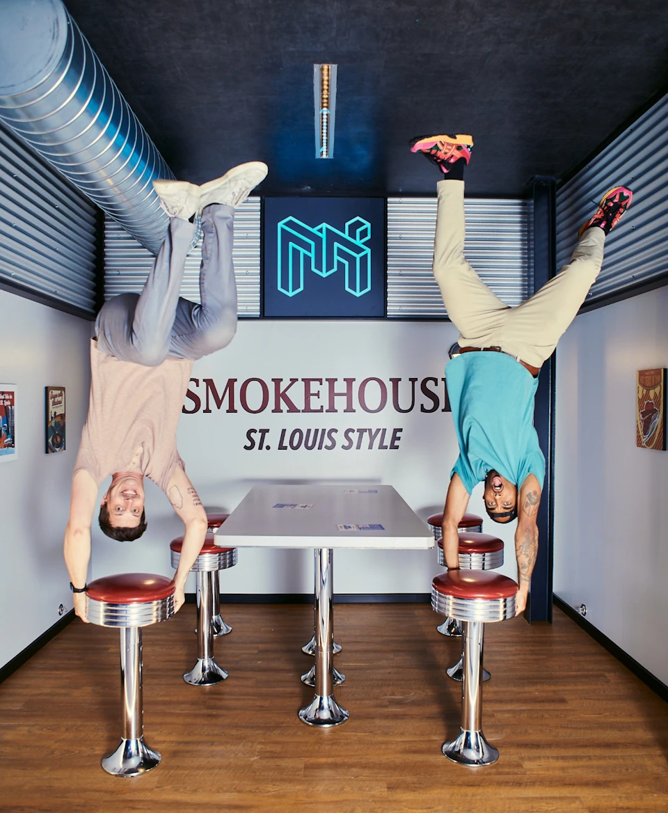 Two individuals are posed upside-down in a retro-style diner setting with a "Smokehouse St. Louis Style" sign in the background.