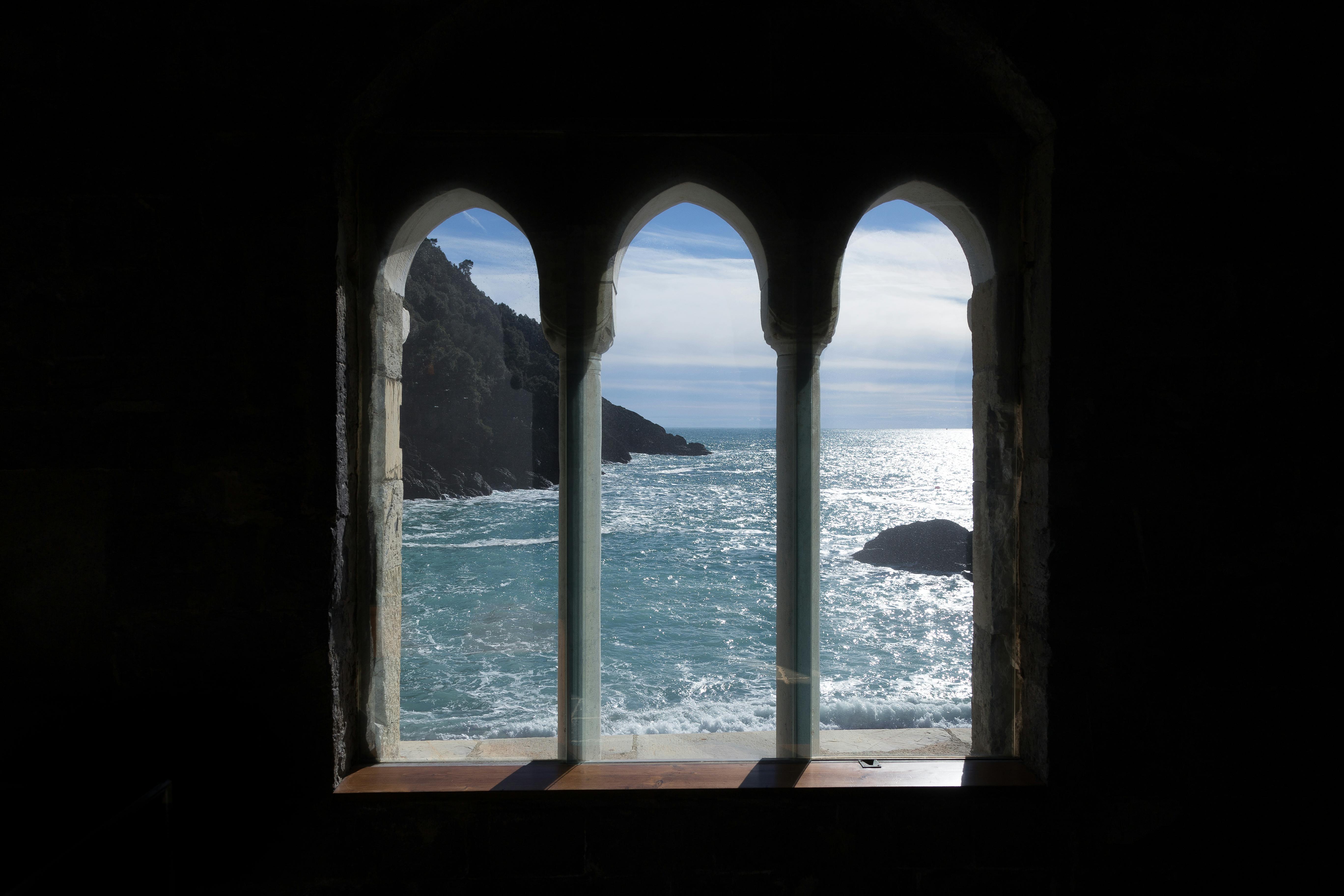 透过石墙上的三扇拱形窗户，可以看到大海和悬崖，阳光反射在水面上。