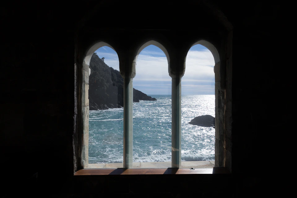 A view of the ocean and cliffs through three arched windows in a stone wall, with sunlight reflecting on the water.