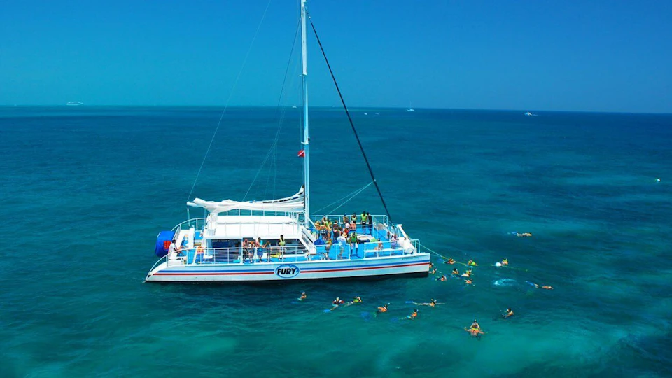 A catamaran named "Fury" floats in clear blue waters surrounded by people snorkeling nearby.