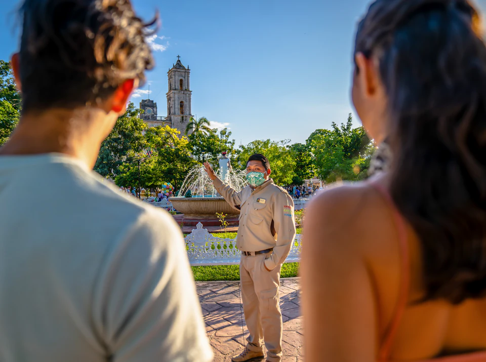 A tour guide wearing a mask speaks to two people near a park fountain with a historic building in the background.