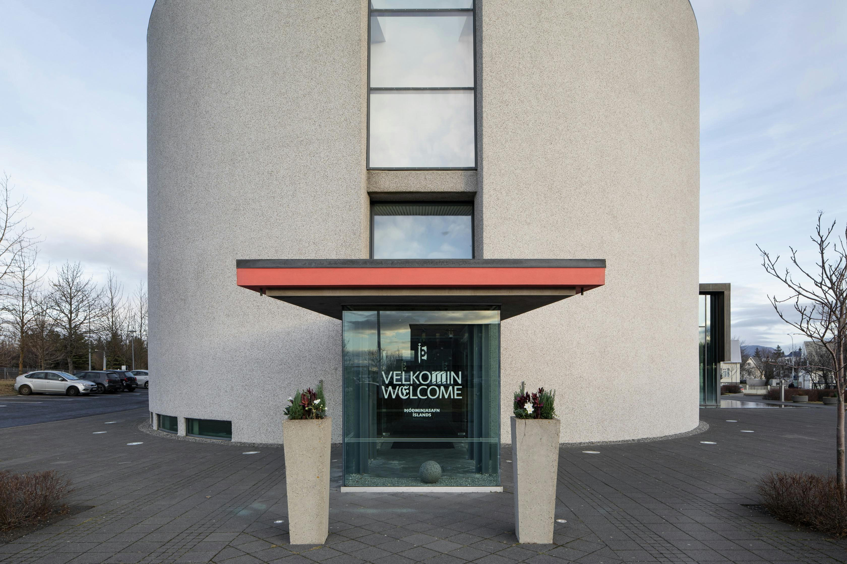 Runder Gebäudeeingang mit "Welcome"-Schild, flankiert von zwei hohen Pflanzkübeln. Im Hintergrund sind ein Parkplatz und Bäume zu sehen.