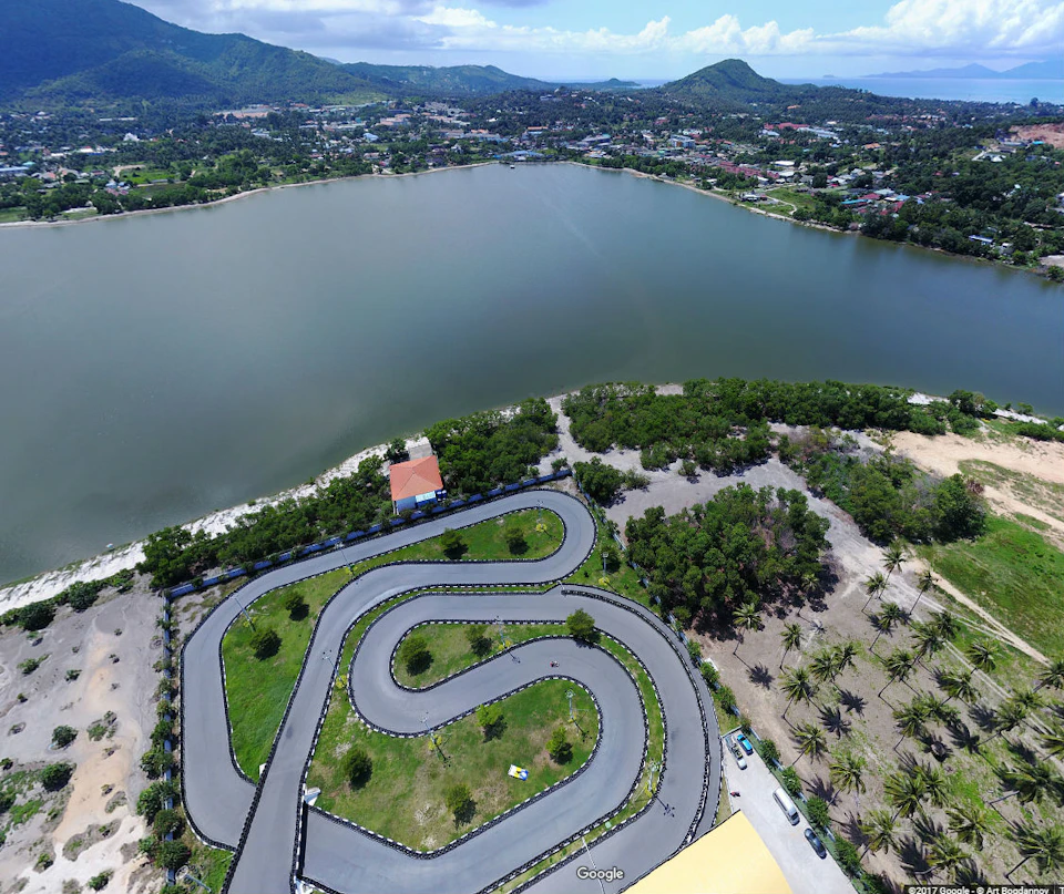 Winding go-kart track surrounded by greenery and a lake, with a distant view of mountains and a town.