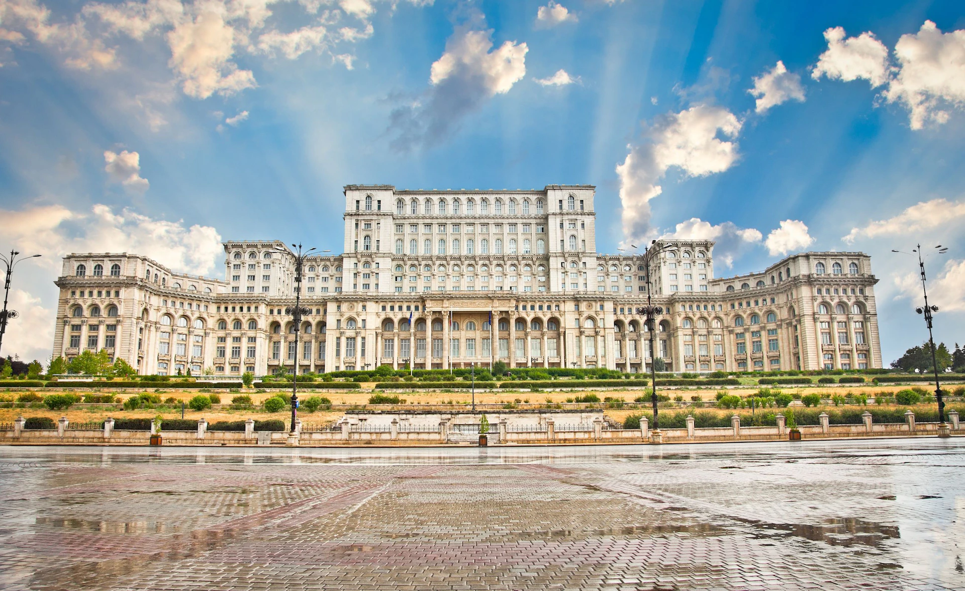 Large neoclassical building with numerous windows and arches, set against a blue sky with clouds, and a wet pavement foreground.