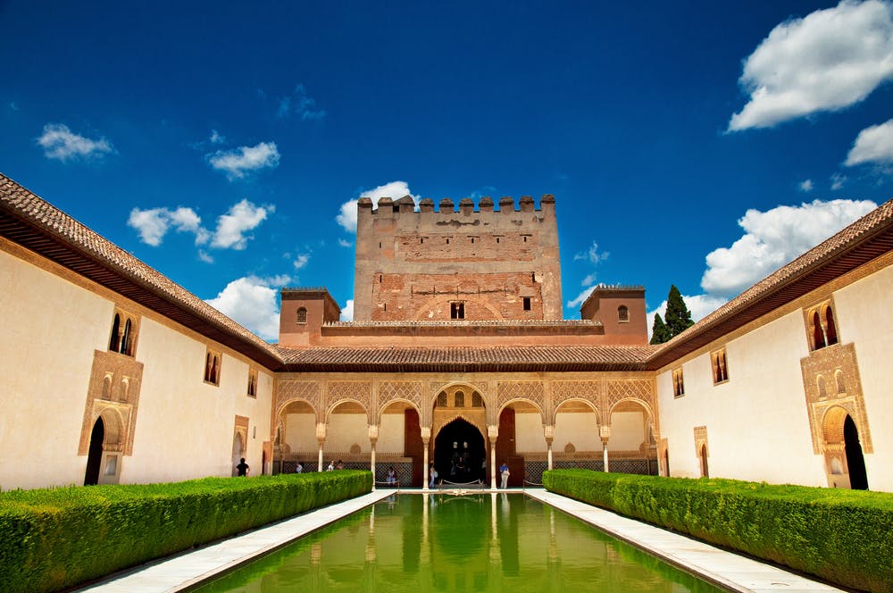 A historical palace courtyard with arched walkways, neatly trimmed hedges, and a reflective pool under a bright blue sky.