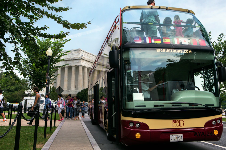 A sightseeing double-decker bus with passengers on the upper deck, parked near an iconic columned building surrounded by trees.